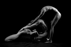 Three nude figures in a linked pose, one bending over two others, in black and white against a black background