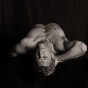 Nude figure lying on a dark surface, photographed from above so the face appears upside down, looking directly into the camera
