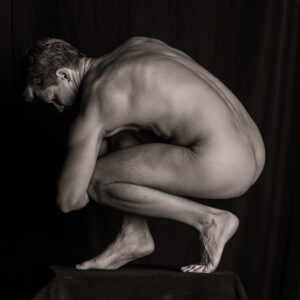 Nude figure crouched on the balls of the feet on a draped box, seen in profile with the head bowed toward the knees.