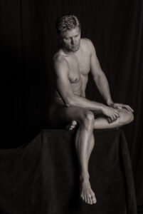 Seated nude on a draped box, torso turned and gaze lowered, lit from the side against a dark curtain.