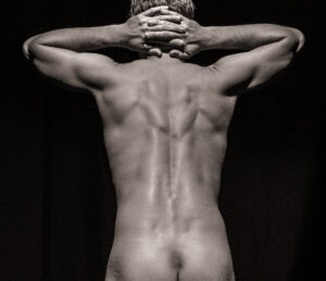 Rear view of a nude figure with both hands clasped behind the head, back muscles shaped by side light
