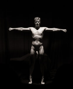 Standing nude figure facing the camera with arms extended straight out to the sides, lit against darkness.