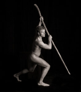 Nude figure in a deep lunge holding a long wooden staff overhead, gaze fixed along its length.